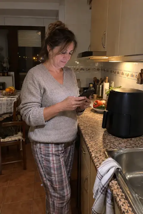 Mujer en pijama mirando el móvil por la noche en su cocina, junto a la freidora de aire